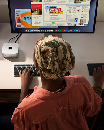 Una persona seduta a una scrivania che usa una Magic Keyboard e un Magic Trackpad per lavorare con un Mac mini collegato a uno Studio Display, accanto ci sono altri oggetti