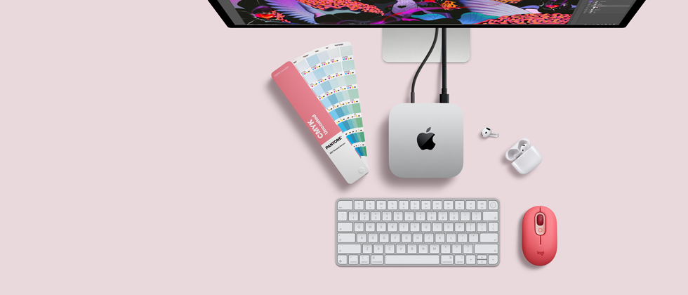 Un Mac mini visto dall’alto, alluminio color argento, angoli arrotondati, logo Apple nero lucido al centro, collegato via cavo a Studio Display parzialmente visibile in alto, AirPods 4 con custodia aperta, campioni di colore Pantone, tastiera e mouse