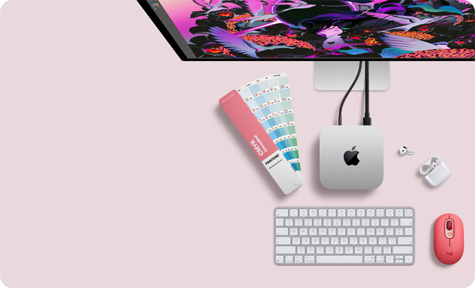 Un Mac mini visto dall’alto, alluminio color argento, angoli arrotondati, logo Apple nero lucido al centro, collegato via cavo a Studio Display parzialmente visibile in alto, AirPods 4 con custodia aperta, campioni di colore Pantone, tastiera e mouse